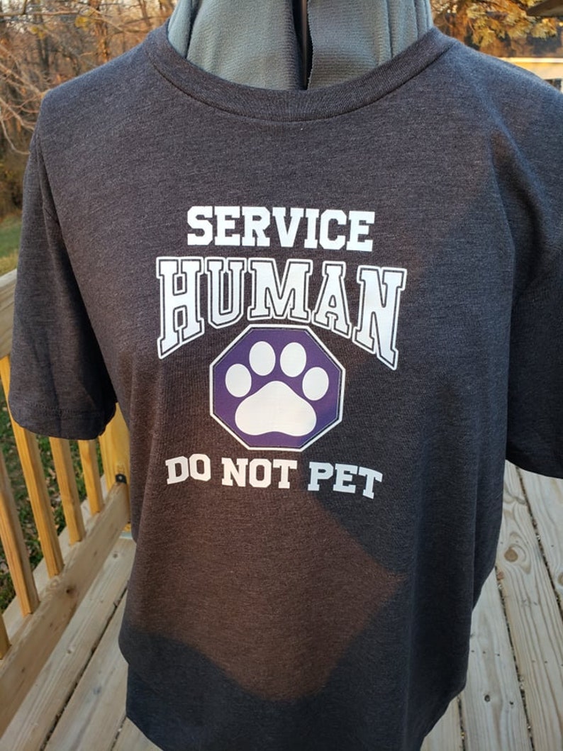 May include: A dark gray t-shirt with the text "SERVICE HUMAN" in white letters and a purple paw print inside a hexagon. The text "DO NOT PET" is below the paw print.