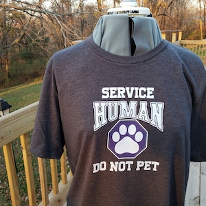 May include: A dark gray t-shirt with white text that reads "SERVICE HUMAN" and a purple paw print with white outline. The text "DO NOT PET" is below the paw print.
