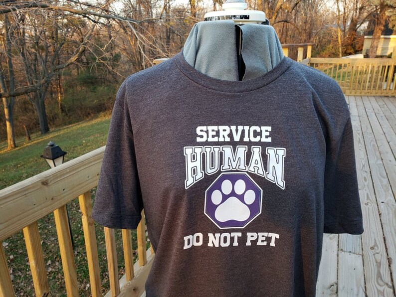 May include: A dark gray t-shirt with white text that reads "SERVICE HUMAN" and a purple paw print with white outline. The text "DO NOT PET" is below the paw print.