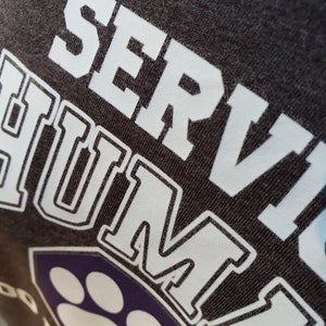 May include: A brown t-shirt with white text that reads "SERVICE HUMANITY" with a purple paw print below the text.