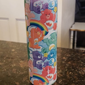 May include: A stainless steel tumbler featuring a colorful design of Care Bears. The design includes various Care Bears characters, rainbows, clouds, and stars. The tumbler has a silver lid and is designed to keep beverages cold or hot.