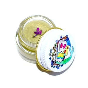 Könnte beinhalten: Ein kleines Glasgefäß mit einem goldenen Deckel und einem weißen Deckel mit einer bunten Illustration eines Cartoon-Aliens mit einem Stern und Blumen. Das Gefäß enthält einen goldenen Glitzer-Lippenbalsam mit einer violetten Blume darin.