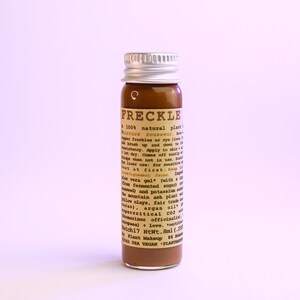 May include: A small brown glass bottle with a silver cap. The bottle contains a brown liquid and has a label with the word "FRECKLE" and other text. The product is a 100% natural plant-based product.