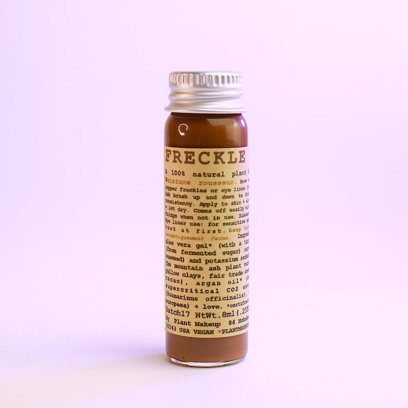 May include: A small brown glass bottle with a silver cap. The bottle contains a brown liquid and has a label with the word "FRECKLE" and other text. The product is a 100% natural plant-based product.