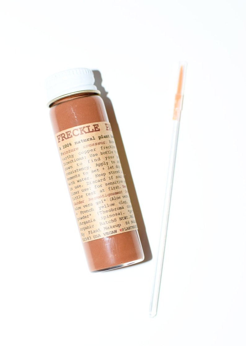 May include: A small glass bottle of "FRECKLE PAINT" with a white cap, filled with a brown powder, and a thin applicator. The label includes text about the product's natural ingredients and use.