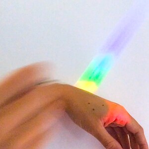 May include: A hand with a rainbow light effect. The light transitions from yellow to green, blue, and purple. The hand is in the foreground, with a white background.