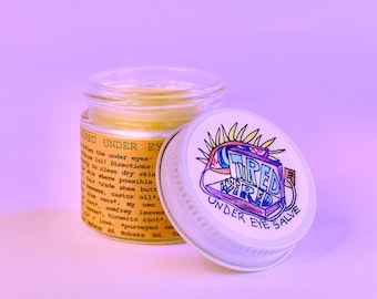 May include: A small glass jar of yellow salve with a white lid. The lid features a colourful graphic with the words "Tired and Wired Under Eye Salve." The jar's label includes text about the product's ingredients and directions.