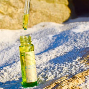 May include: A small, green glass bottle with a yellow liquid and a dropper. The bottle has a label with text. The dropper is suspended above the bottle, with a drop of liquid about to fall. The background is a sandy surface.