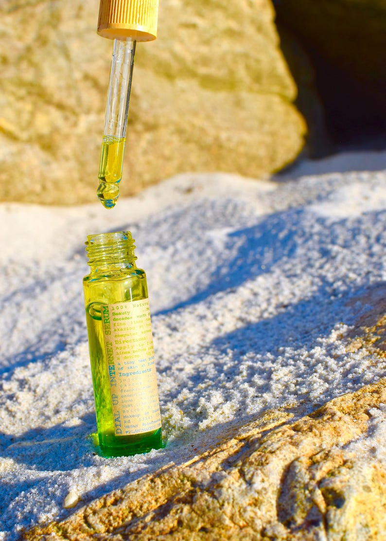 May include: A small, green glass bottle with a yellow liquid and a dropper. The bottle has a label with text. The dropper is suspended above the bottle, with a drop of liquid about to fall. The background is a sandy surface.