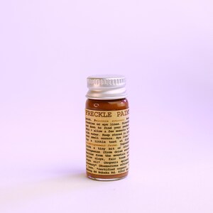 May include: A small glass bottle of "FRECKLE PAIN" with a silver cap. The bottle contains a brown liquid and has a label with text. The product is designed to create freckles or be used on eye lines.