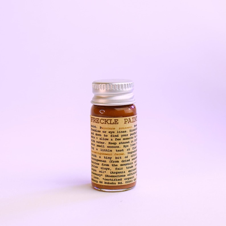 May include: A small glass bottle of "FRECKLE PAIN" with a silver cap. The bottle contains a brown liquid and has a label with text. The product is designed to create freckles or be used on eye lines.