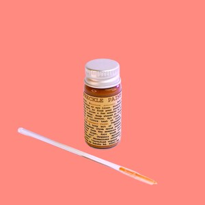 May include: A small glass bottle of freckle paint with a silver lid and a white applicator stick. The bottle has a brown liquid inside and a label with text. The background is a solid coral color.