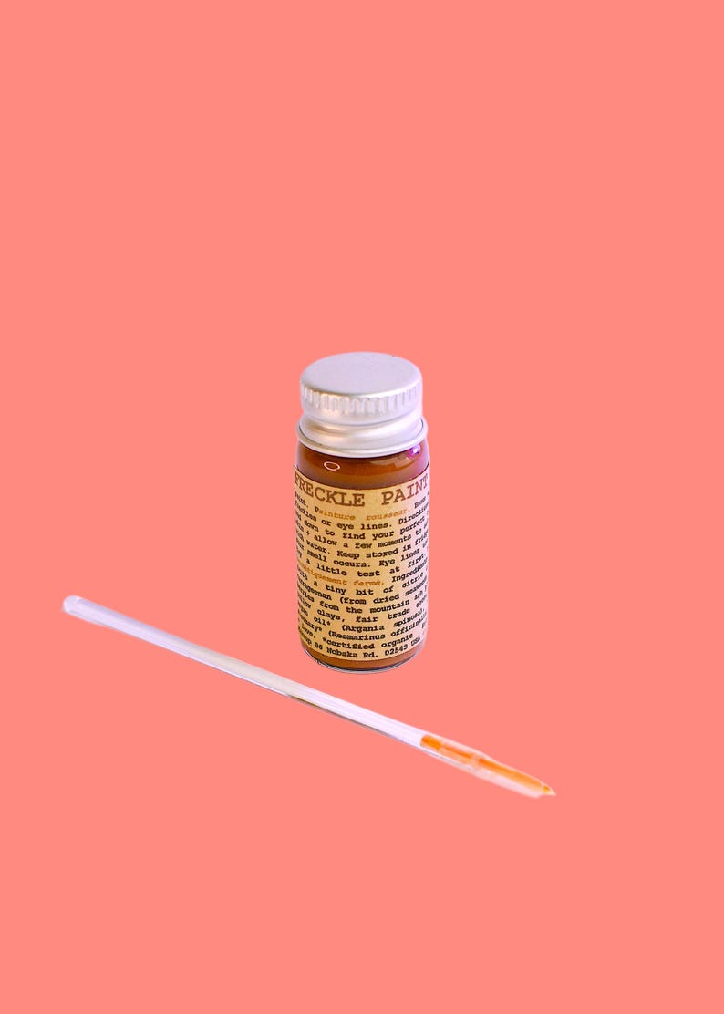 May include: A small glass bottle of freckle paint with a silver lid and a white applicator stick. The bottle has a brown liquid inside and a label with text. The background is a solid coral color.