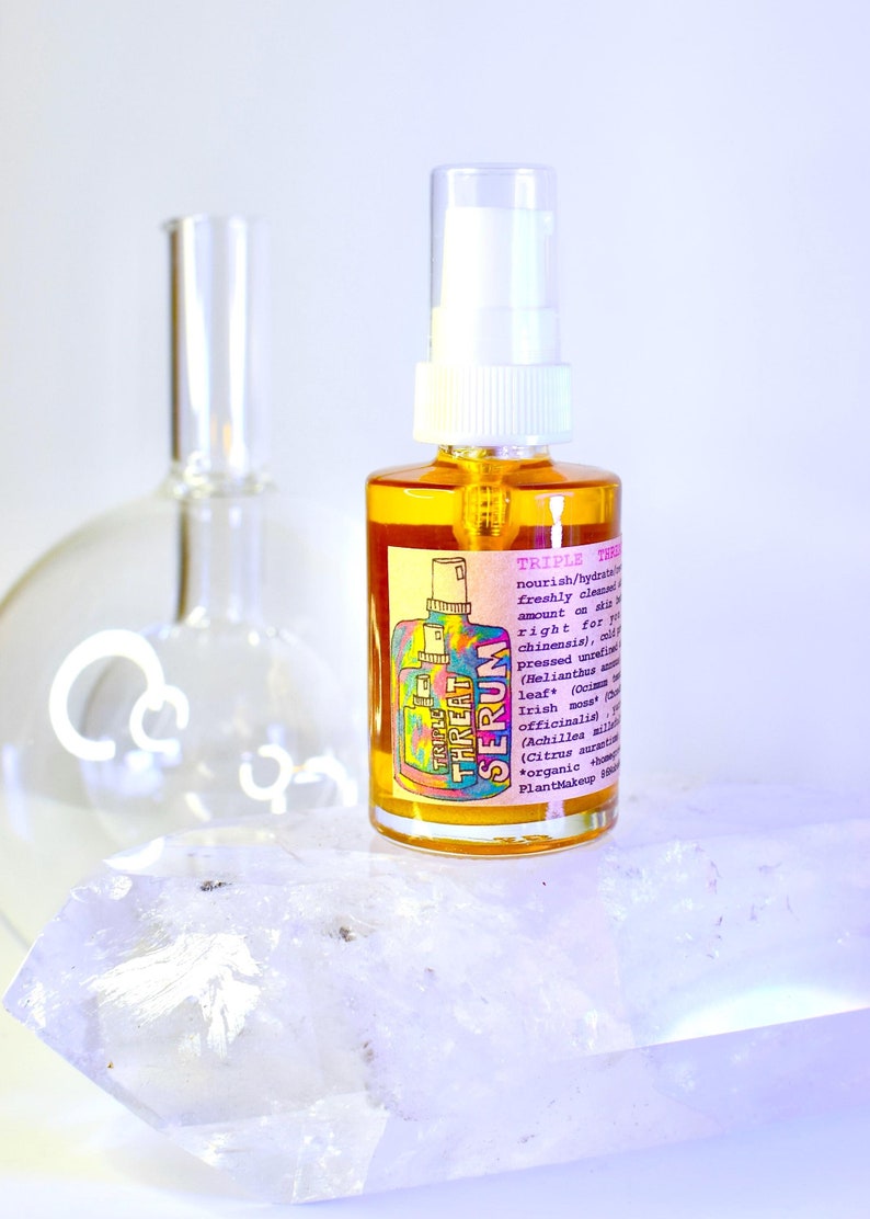 May include: A clear glass bottle with a white spray nozzle, filled with a golden liquid, labeled "Triple Threat Serum." The label features colorful text and a graphic of a bottle. The bottle rests on a clear crystal with a blurred glass flask in the background.