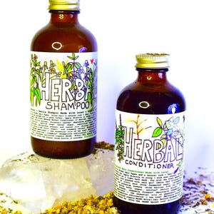 May include: Two amber glass bottles with gold lids, labeled "Herbal Shampoo" and "Herbal Conditioner". Both bottles feature hand-drawn illustrations of herbs and flowers. The bottles are sitting on a white surface with dried herbs and flowers scattered around them.