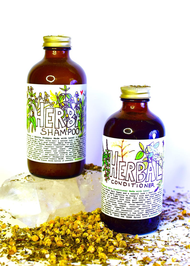 May include: Two amber glass bottles with gold lids, labeled "Herbal Shampoo" and "Herbal Conditioner". Both bottles feature hand-drawn illustrations of herbs and flowers. The bottles are sitting on a white surface with dried herbs and flowers scattered around them.