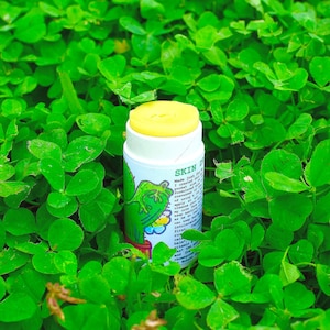 May include: A cylindrical white skin balm stick with a yellow top, featuring a green cactus illustration and text that reads SKIN DEEP. The label includes printed instructions and ingredient information in black text on a white background.