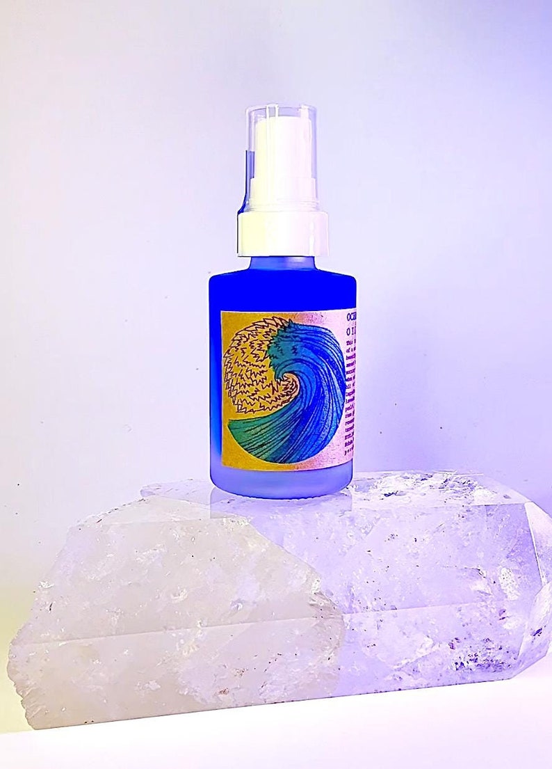 May include: A blue glass bottle with a white pump top and a yellow and blue label with a wave design. The label reads "Ocean Mist" and has a list of ingredients. The bottle is sitting on a white crystal.