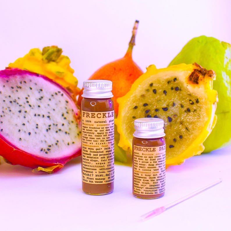 May include: Two small glass bottles with silver caps labeled FRECKLE and FRECKLE PAINT, containing brown liquid. The bottles are surrounded by a sliced pink dragon fruit, a yellow dragon fruit, and an orange fruit. A small applicator is included.