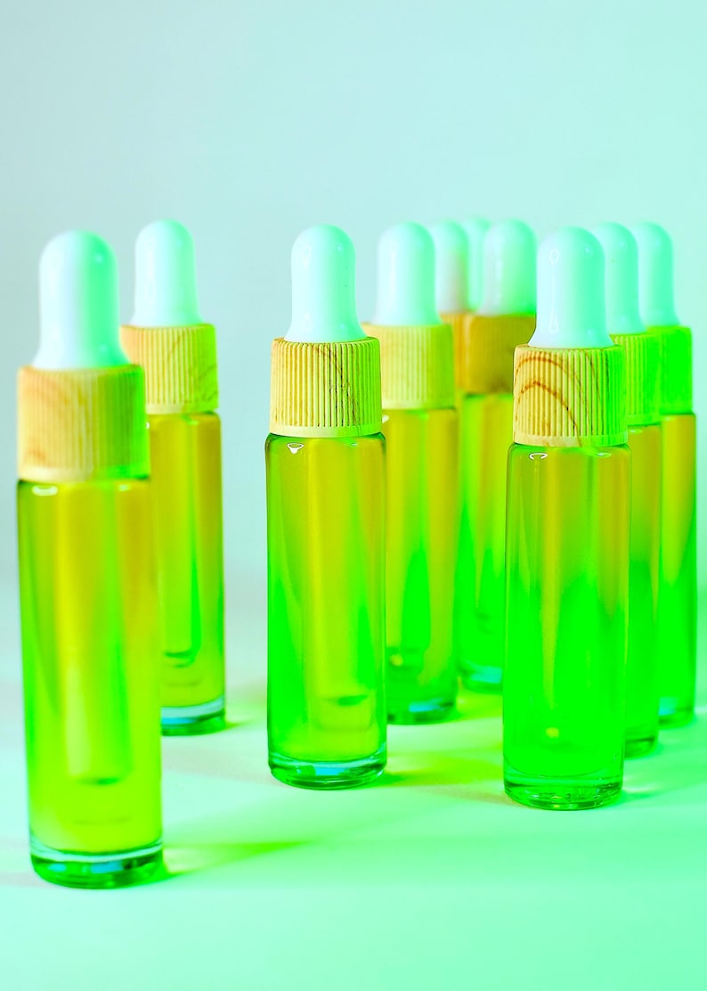 May include: A collection of small glass dropper bottles filled with a vibrant green liquid. Each bottle has a white dropper top and a wooden collar. The bottles are arranged on a light green surface.