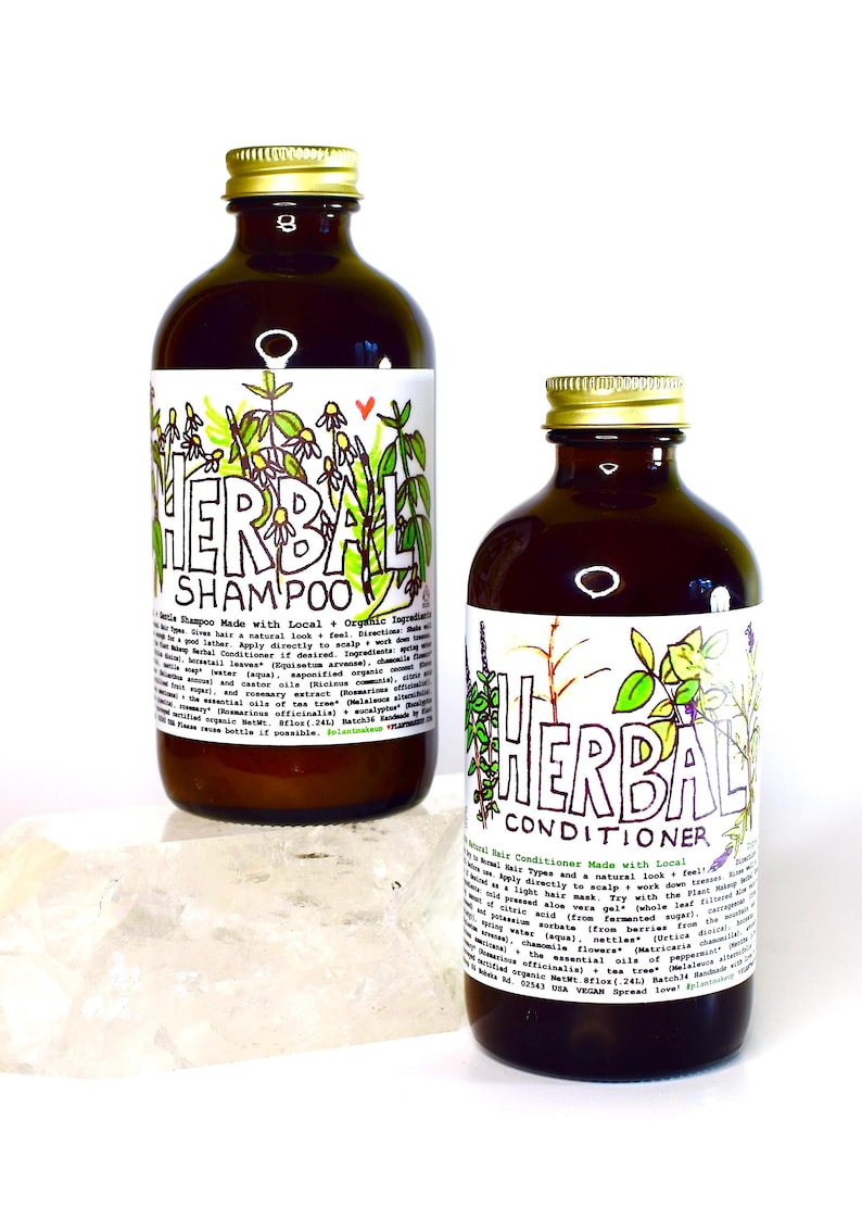 May include: Two brown glass bottles with gold lids, labeled "Herbal Shampoo" and "Herbal Conditioner". Both bottles feature hand-drawn illustrations of plants and flowers. The bottles are sitting on a white surface.