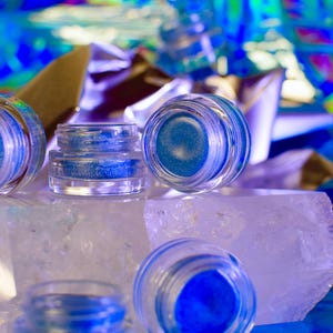 Pu&ograve; includere: Quattro piccoli barattoli di vetro con glitter blu all'interno, appoggiati su una roccia di cristallo bianco. I barattoli sono trasparenti e hanno un coperchio argentato.