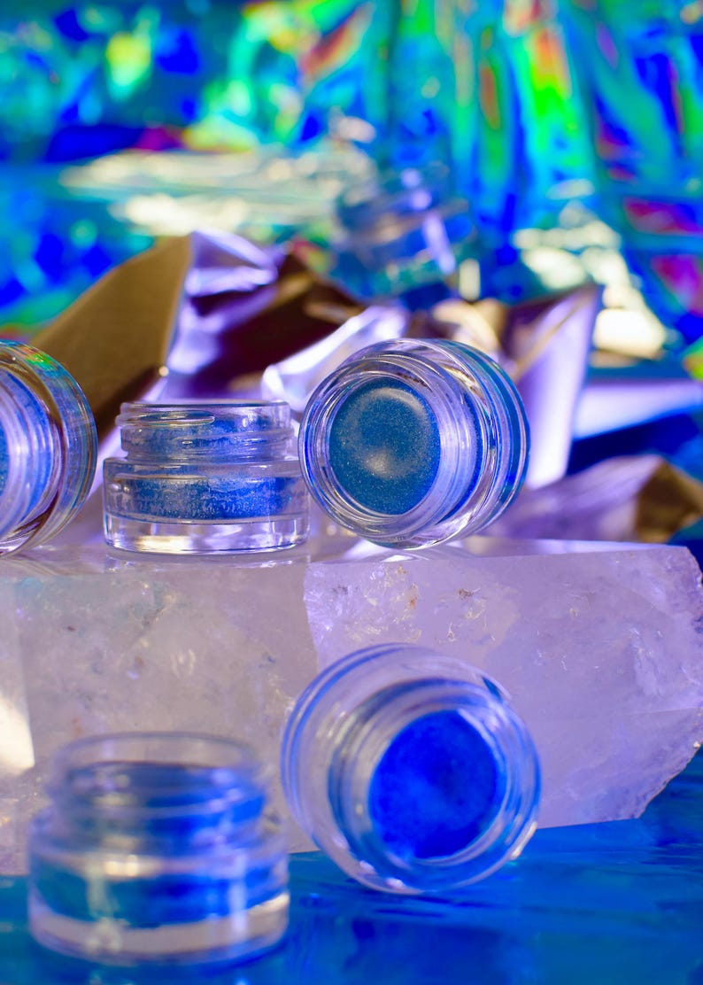 Pu&ograve; includere: Quattro piccoli barattoli di vetro con glitter blu all'interno, appoggiati su una roccia di cristallo bianco. I barattoli sono trasparenti e hanno un coperchio argentato.