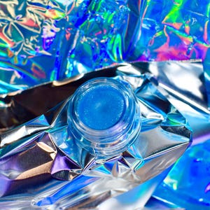 Pu&ograve; includere: Un piccolo barattolo di lucidalabbra blu con una finitura scintillante. Il barattolo &egrave; appoggiato su un pezzo di foglio di alluminio iridescente.