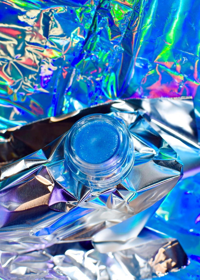 Pu&ograve; includere: Un piccolo barattolo di lucidalabbra blu con una finitura scintillante. Il barattolo &egrave; appoggiato su un pezzo di foglio di alluminio iridescente.
