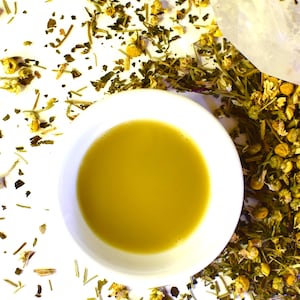 May include: A white ceramic cup filled with a golden yellow herbal tea. The tea is surrounded by loose dried herbs and flowers on a white background.