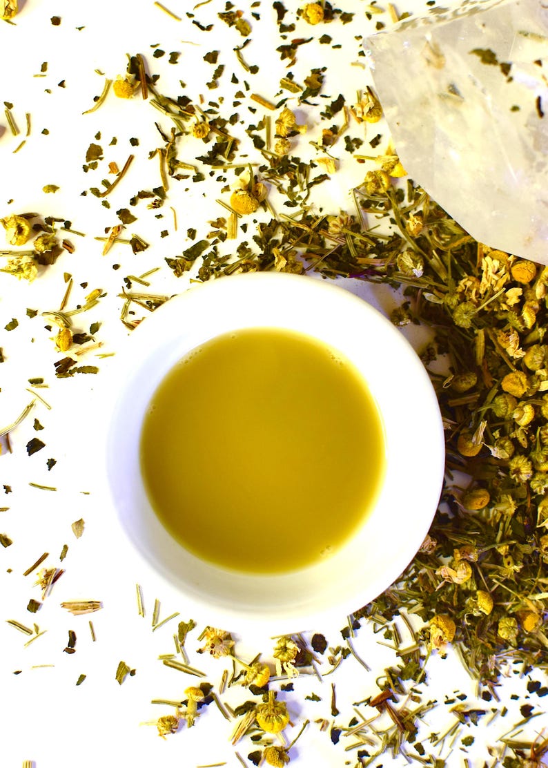 May include: A white ceramic cup filled with a golden yellow herbal tea. The tea is surrounded by loose dried herbs and flowers on a white background.