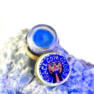 Pu&ograve; includere: Un piccolo barattolo di gloss per labbra blu glitterato con un tappo dorato. Il tappo ha un adesivo con un viso di cartone animato con stelle intorno agli occhi e le parole "Space Goth Gloss".