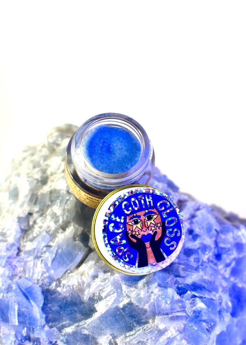Pu&ograve; includere: Un piccolo barattolo di gloss per labbra blu glitterato con un tappo dorato. Il tappo ha un adesivo con un viso di cartone animato con stelle intorno agli occhi e le parole "Space Goth Gloss".