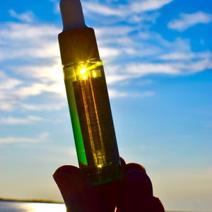 May include: A clear glass bottle with a white dropper top, filled with a green liquid. The bottle is held up against a bright blue sky with the sun shining through it. The bottle is held in a hand.