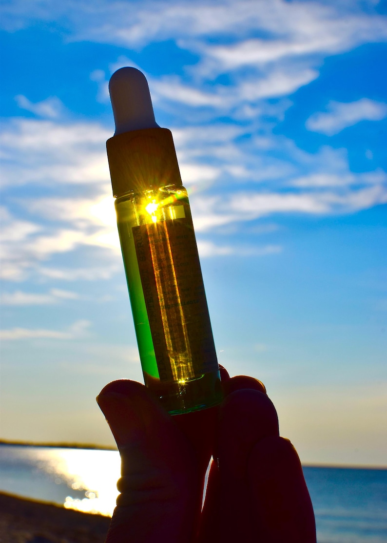 May include: A clear glass bottle with a white dropper top, filled with a green liquid. The bottle is held up against a bright blue sky with the sun shining through it. The bottle is held in a hand.