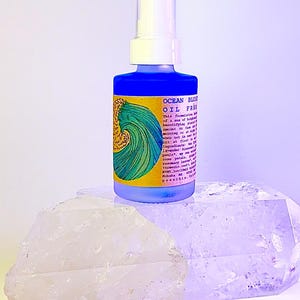 May include: A blue glass bottle with a white pump top and a yellow label that says "OCEAN BLOSSOM OIL FREE" and lists ingredients. The bottle is sitting on a white crystal.
