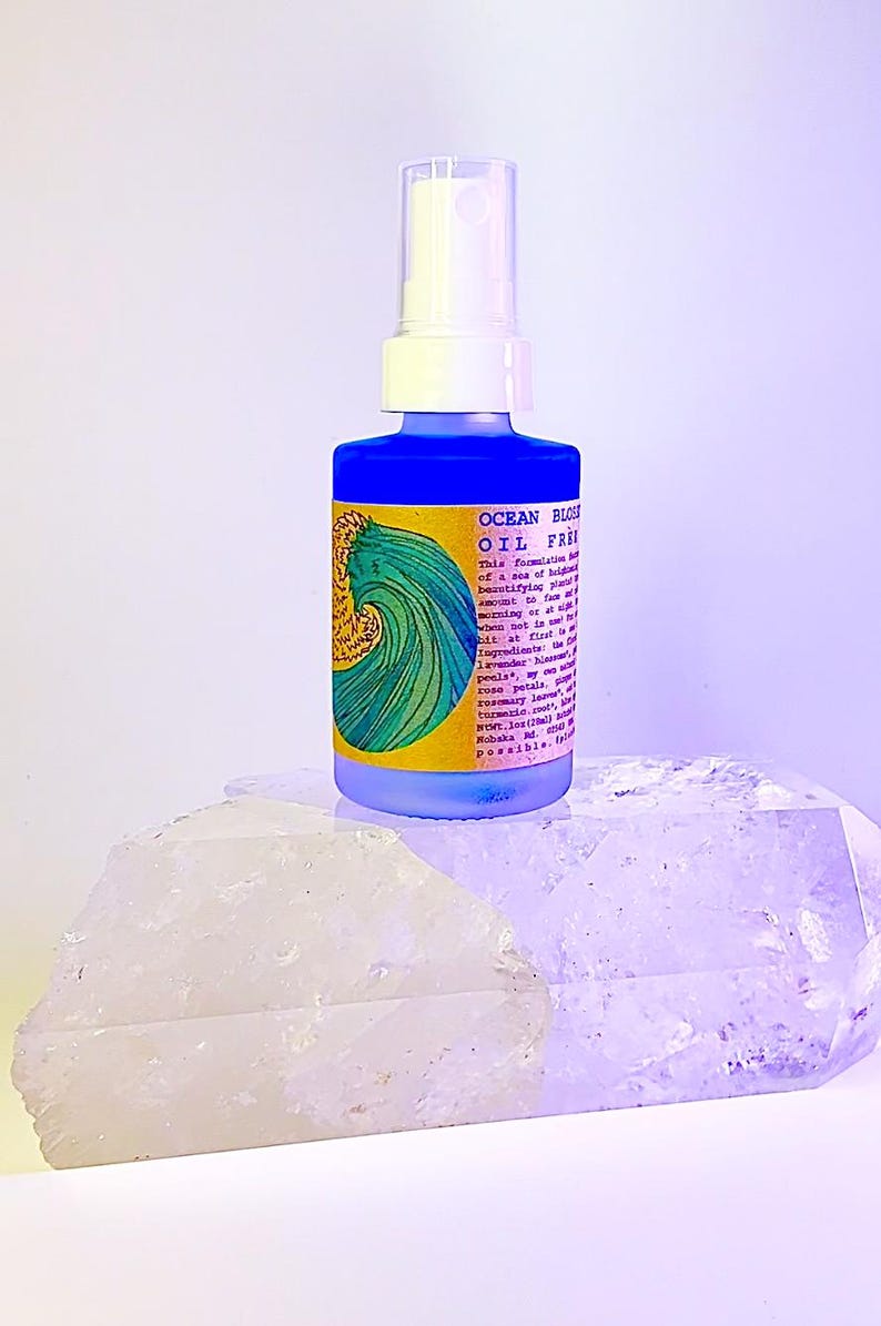 May include: A blue glass bottle with a white pump top and a yellow label that says "OCEAN BLOSSOM OIL FREE" and lists ingredients. The bottle is sitting on a white crystal.