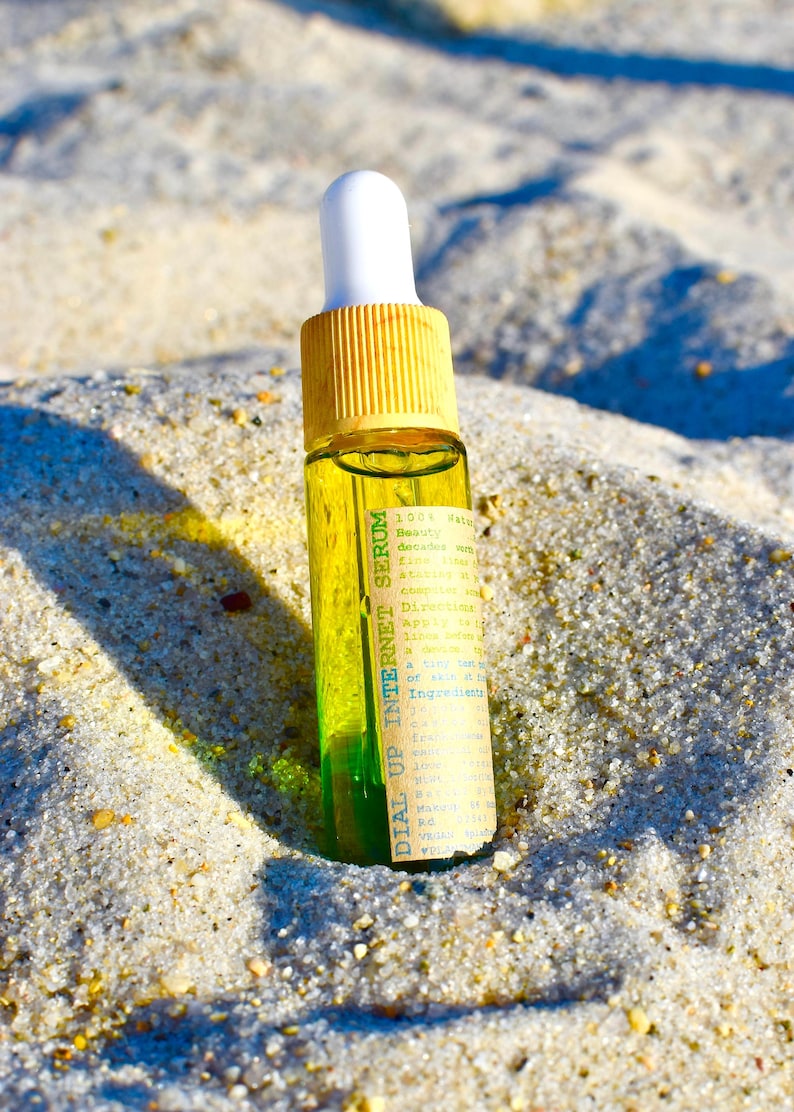 May include: A small glass bottle of serum with a yellow cap and white dropper. The bottle is filled with a clear liquid and a green liquid at the bottom. The label reads "DIAL UP INTERNET SERUM". The bottle is resting on sand.