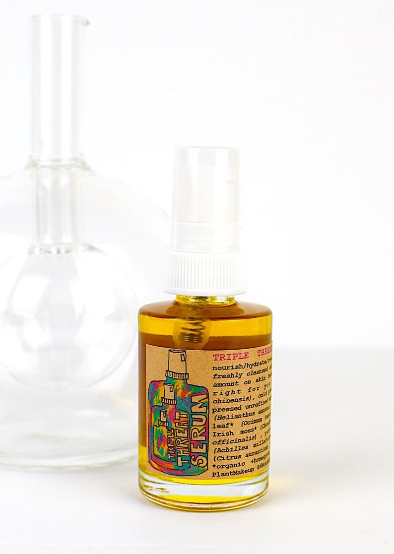 May include: A clear glass bottle with a white spray top, filled with a golden liquid. The label reads "Triple Threat Serum" with a colorful graphic. A larger, clear glass bottle is in the background.