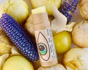 May include: A cylindrical cardboard tube of eye balm labeled i u under eye balm with a hand-drawn eye illustration, surrounded by several yellow husked tomatillos and two ears of small, deep purple ornamental corn with glossy kernels.