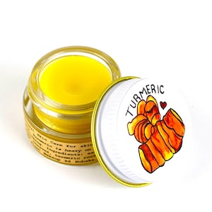 May include: A small glass jar with a yellow lid and a white label that says "Skin Care For skin Turmeric is heavy on Ingredients: ... turmeric root ... Makeup 86 Hobaka". The jar is open and the lid is on the side. The lid has a white background with a drawing of a piece of turmeric root and the word "TURMERIC" in black letters.