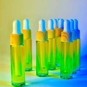 May include: A collection of clear glass dropper bottles filled with a vibrant green liquid. Each bottle has a white and blue dropper top with a yellow wooden collar. The bottles are arranged on a gradient background of yellow and blue.