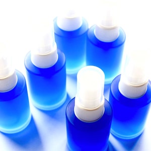 May include: Six small blue spray bottles with white tops. The bottles are arranged in a circle on a white surface.