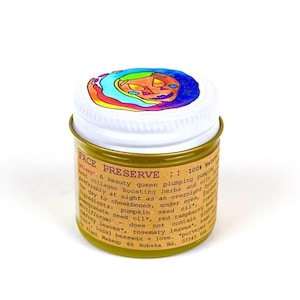 Può includere: Un piccolo barattolo rotondo del prodotto per la cura della pelle Face Preserve. Il barattolo ha una base color oro e un coperchio bianco con un'illustrazione colorata. L'etichetta include testo sugli ingredienti e l'uso del prodotto.