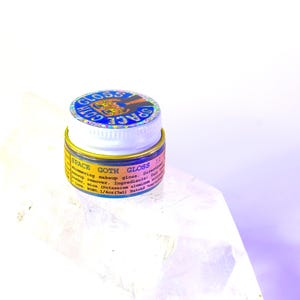 Pu&ograve; includere: Un piccolo barattolo rotondo di gloss per il trucco Space Goth Gloss. Il barattolo &egrave; blu con un coperchio dorato e un adesivo olografico con le parole "Space Goth Gloss" su di esso. Il barattolo &egrave; su una superficie bianca.