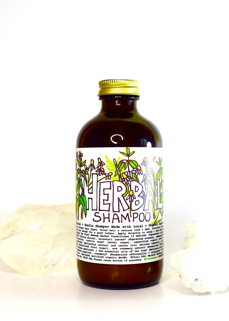 May include: A brown glass bottle with a gold lid, labeled "Herbal Shampoo". The bottle is surrounded by a white background and a white crystal.