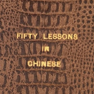 Puede incluir: Una portada de libro marrón con un patrón texturizado similar al de un cocodrilo. Letras doradas deletrean "FIFTY LESSONS IN CHINESE" y el nombre del autor, "CHANG". El diseño del libro sugiere un enfoque en el aprendizaje de idiomas.