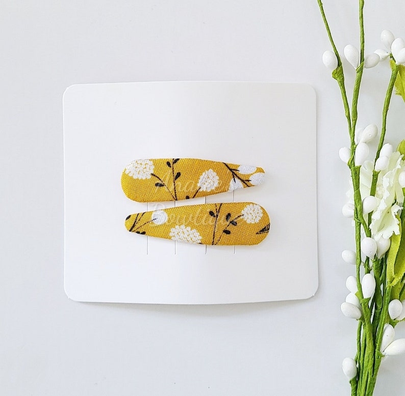 May include: Two fabric-covered hair clips featuring a mustard yellow background with a white dandelion and black stem floral pattern. The clips are attached to a rectangular white display card, positioned horizontally one above the other.