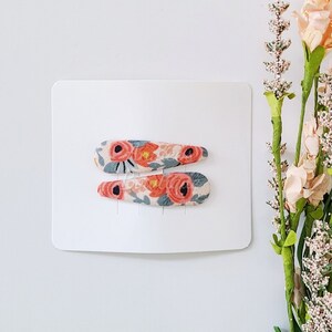 May include: Two hair clips featuring a fabric covering with a floral pattern in shades of coral, peach, and muted blue on a cream background. The clips are attached to a rectangular white display card, positioned horizontally one above the other.