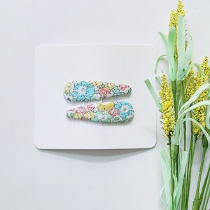 May include: Two teardrop-shaped hair clips featuring a white base with a colorful floral pattern in shades of light blue, yellow, and pink. The hair clips are attached to a rectangular white display card, positioned horizontally one above the other.
